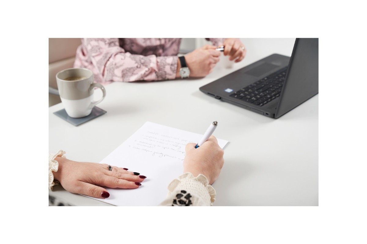 A person's hands are shown writing with a pen on a white piece of paper, next to a laptop on a white desk.