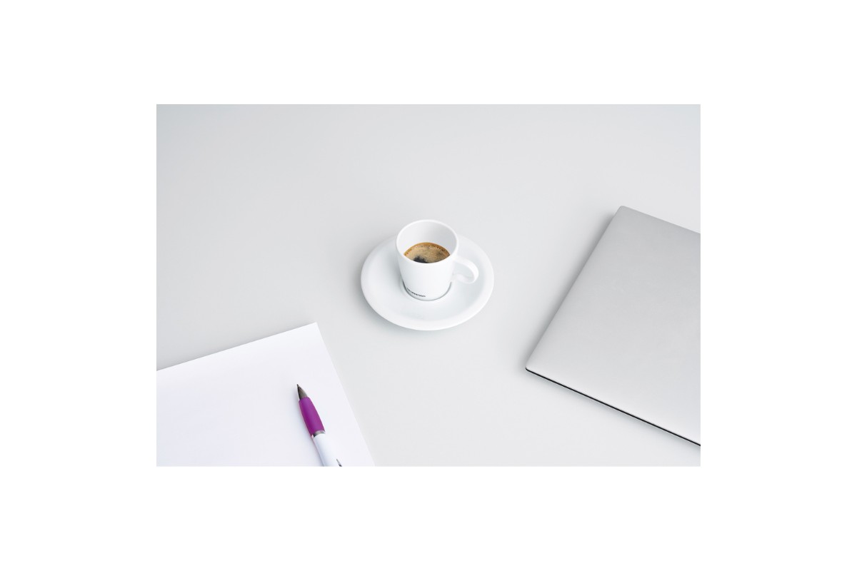 An overhead shot of a light grey desk with a white notepad and a purple pen on the left, a white espresso cup and saucer in the centre, and a silver laptop on the right.