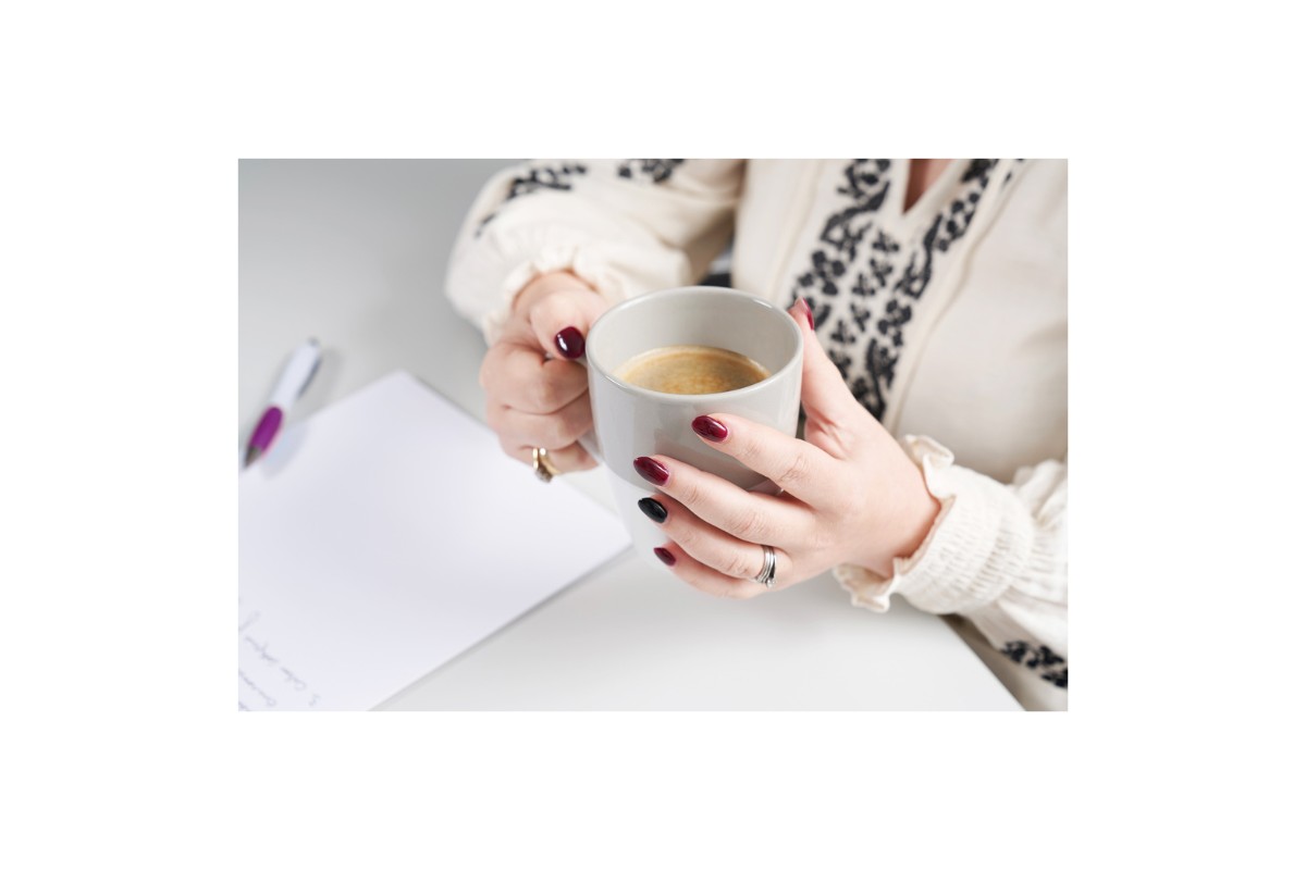 A close-up of a person's hands holding a gray mug of coffee. The person has dark red and black nail polish. A pen and paper are also on the desk.