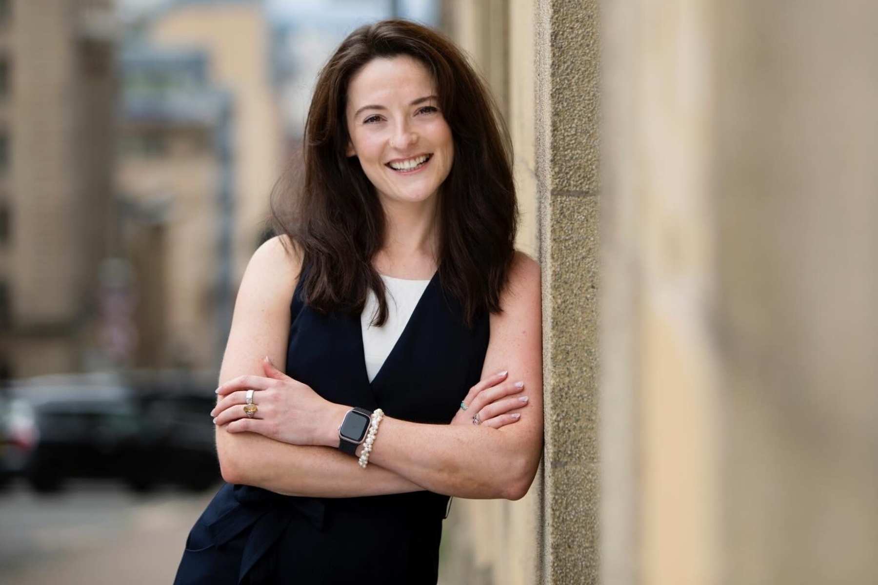Nicole A smiling woman with dark brown hair, wearing a dark sleeveless top with a white v-neck insert, leans against a textured stone wall with her arms crossed.