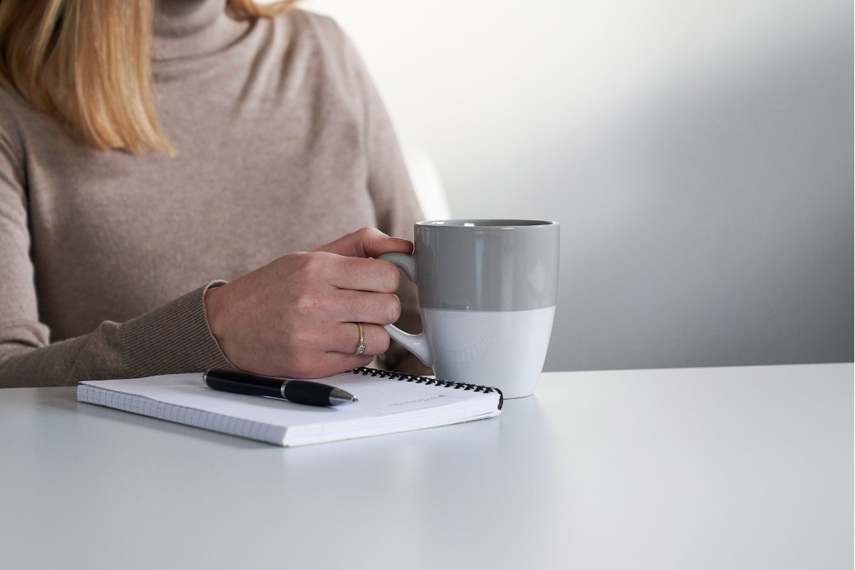 A woman sits at a white table, holding a two-toned mug, with a pen resting on a spiral notebook.