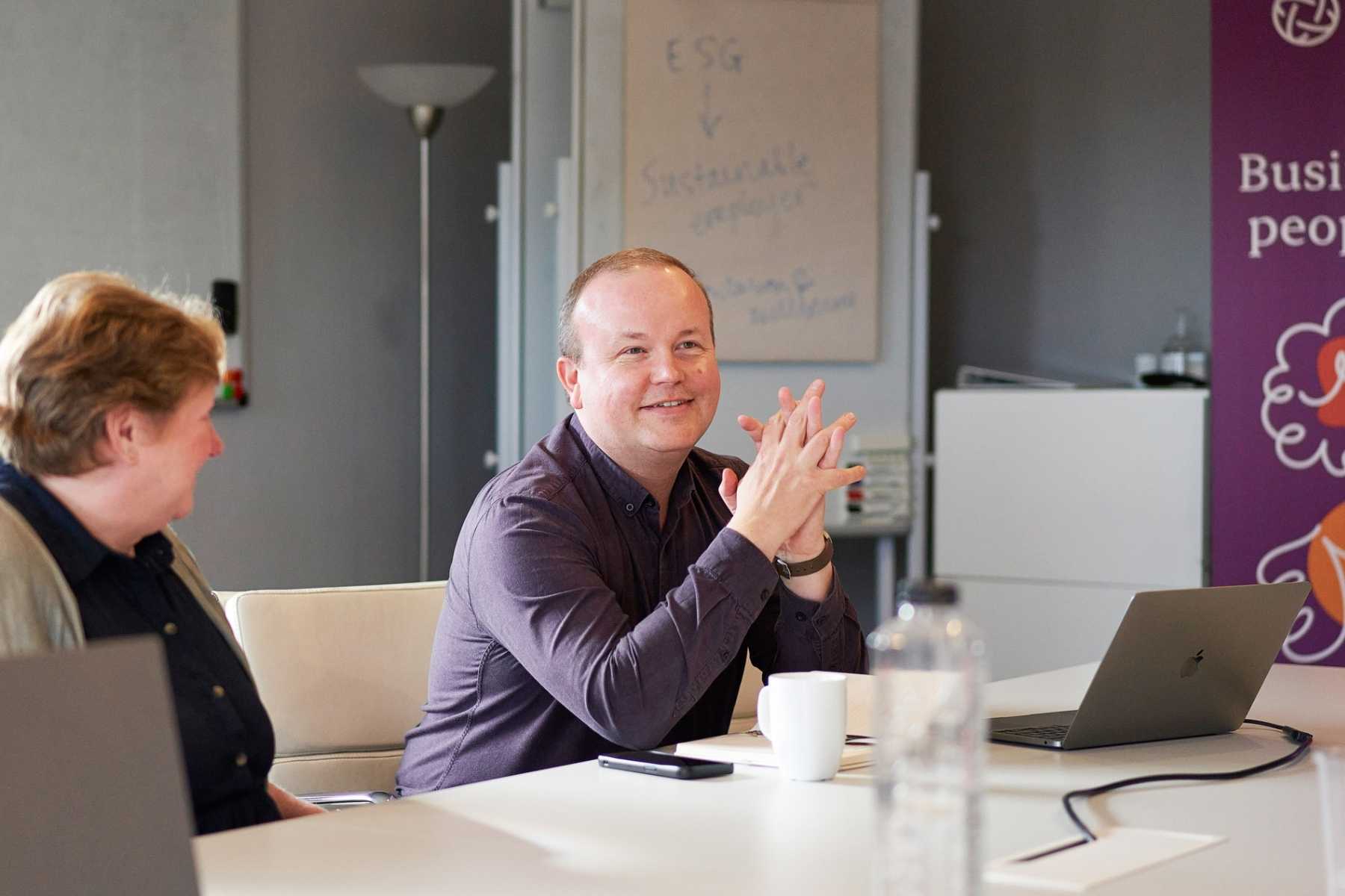 A man in a dark shirt sits at a table, smiling with his hands clasped. A whiteboard with "ESG" written on it is in the background. Another person is seated to the man's left, and a laptop, mug, and water bottle are on the table.